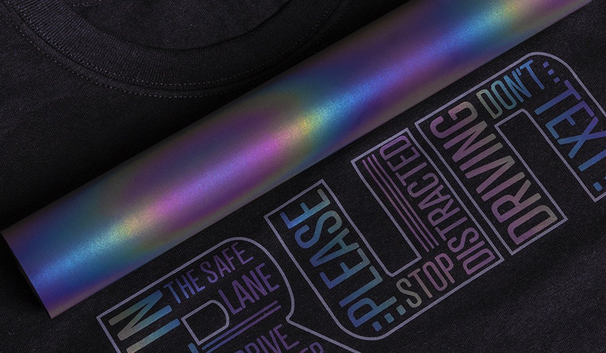 Reflecterende flexfolie: De complete gids voor 2026 3 Een rol iriserende flexfolie ligt op een zwart T-shirt met een reflecterend tekstontwerp.