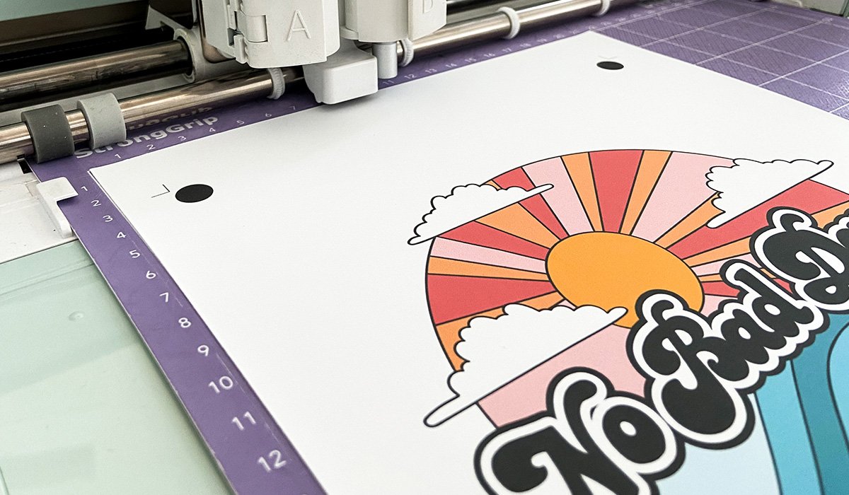Welke printbare flexfolie past het beste bij jouw project? 3 Een wit vel Siser Easy Color Flex met kleurrijke "No Bad Days" print en registratiemarkeringen wordt ingevoerd in een snijmachine.